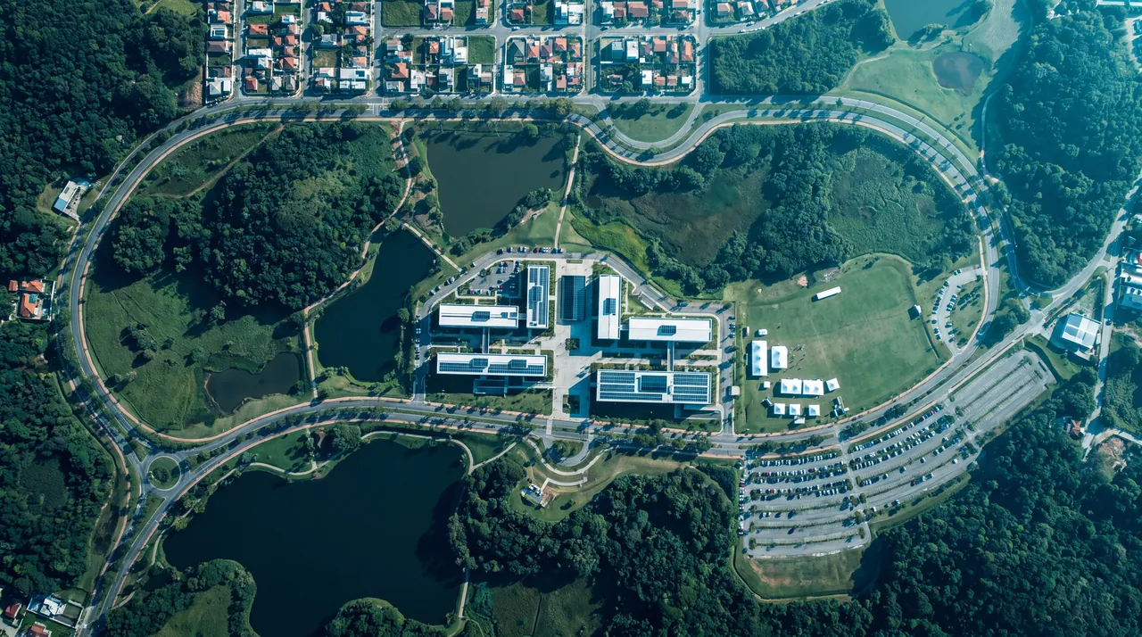 Imagem de satélite conceitual do Sapiens Parque, vista de cima: anel viário cercado de mata, lagos, campus de prédios com telhados solares, campo de tendas ao lado da avenida principal e estacionamento, com o traçado ilustrativo da corrida desenhado sobre as ruas.